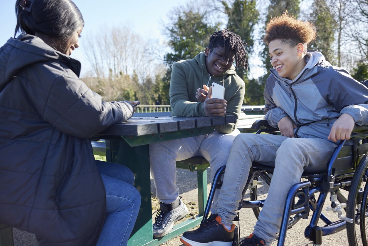 Three people sit at a bench in a park. One person is sitting in a wheelchair while their friend shows them an image on his phone. Another friend sits opposite, smiling at her phone.