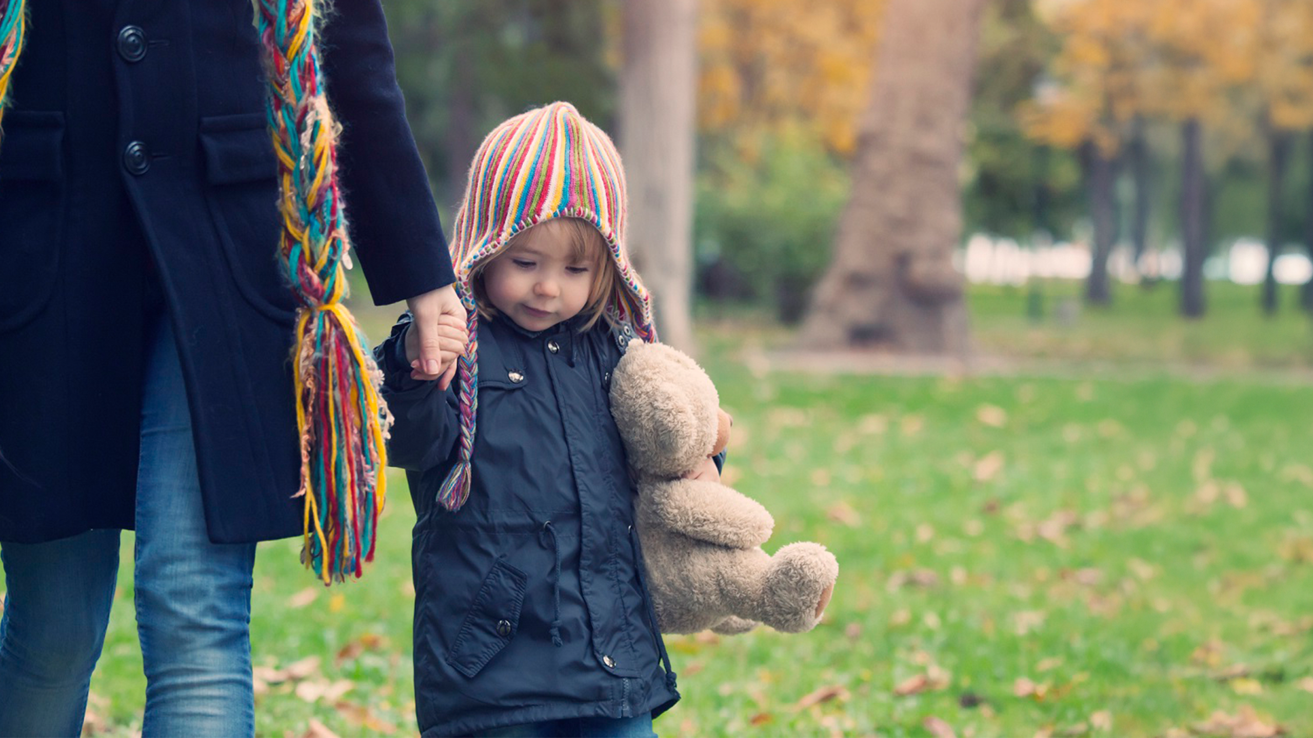 A young girl wearing a stripey, multi-colour hat and navy coat is holding her mother's hand who is mostly out of shot. She is hugging a light brown teddy bear. They are walking in a park with orange and gold-leaved trees.